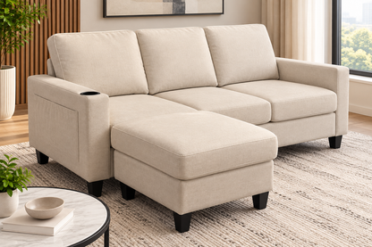 A light beige fabric sectional sofa with a chaise ottoman section, shown in a modern living room setting with a patterned rug, a small round table, and a window.