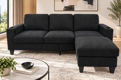 A modern black fabric sectional sofa with a right-facing chaise lounge, shown in a styled living room setting with a round coffee table, potted plants, and a light-colored rug.